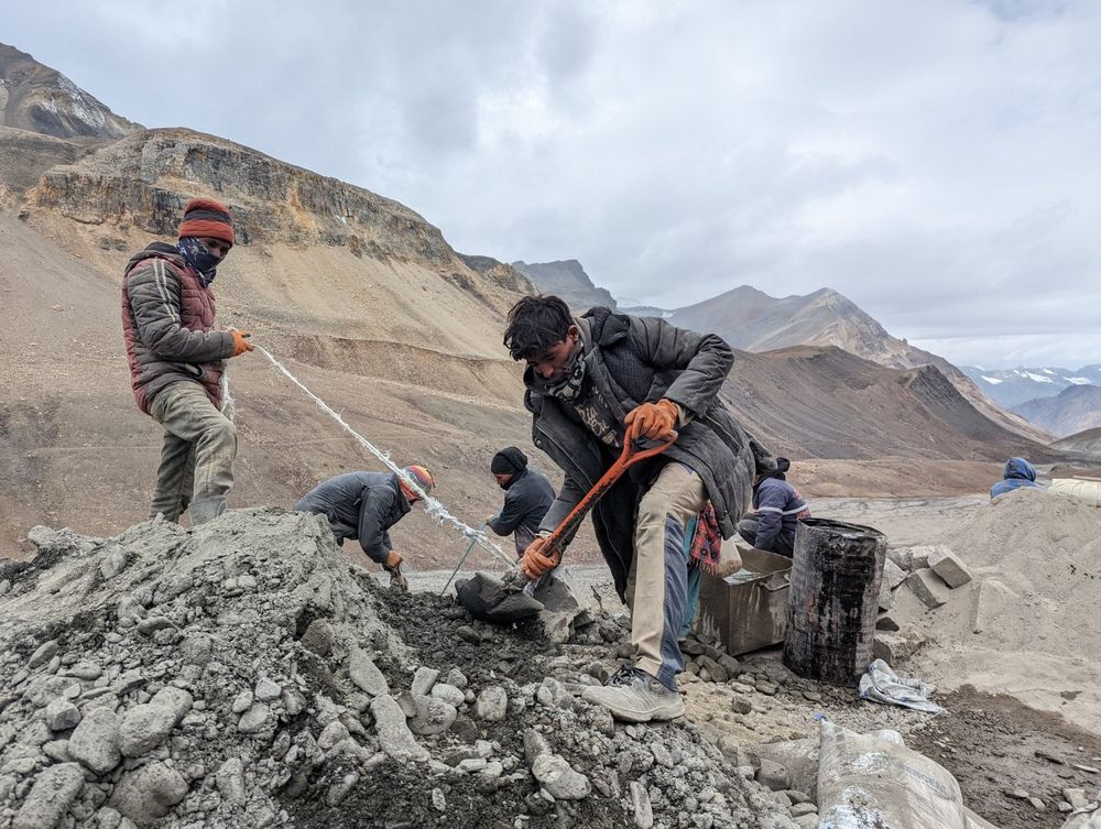 2 Straßenarbeiter räumen mit 1 Schaufel einen Schutthaufen ab. Einer hält eine Metallschaufel, ein anderer zieht helfend mit einem Seil an der Schaufel. Im Hintergrund weitere Arbeiter und eine Feuertonne zum Wärmen, wenn es zu kalt wird.
Die Umgebung ist vegetationslos, im Hintergrund sieht man hohe Berge mit Schnee, wahrscheinlich Gletscher. 
Die Arbeiter sind dick bekleidet: alles deutet auf eine große Höhe hin, in der das Hantieren mit einer Schaufel ohne Unterstützung ein Problem ist. 
Der Himmel ist stark bewölkt. 
