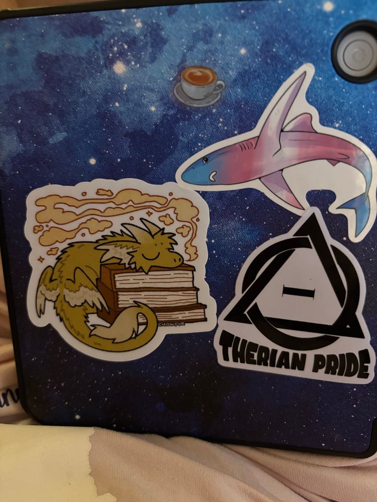 Back of our e reader case with a chow fur book dragon sticker a trans shark sticker a coffee sticker from flam craft and a therian pride sticker by caustic canine 