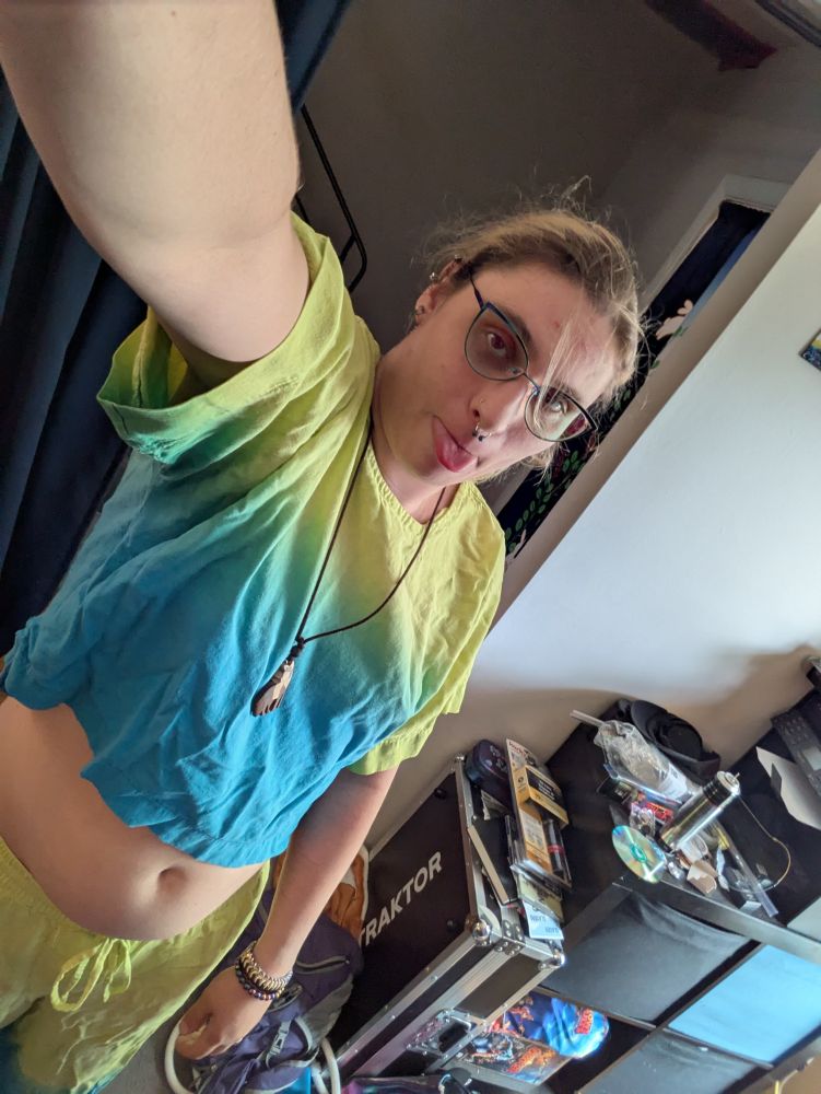 Us in a yellow and blue crop top blepping at the camera with our wooden wolf necklace on 