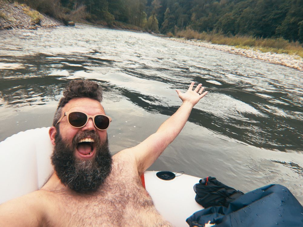 A close-up selfie of a man with a large beard and sunglasses floating on an inner tube. He's smiling widely, with his mouth open, and has one arm raised in the air as if in celebration. The background is a river with gentle rapids and a grassy bank. He seems blissfully unaware that in four hours he will shatter his backside on a giant river stone. 