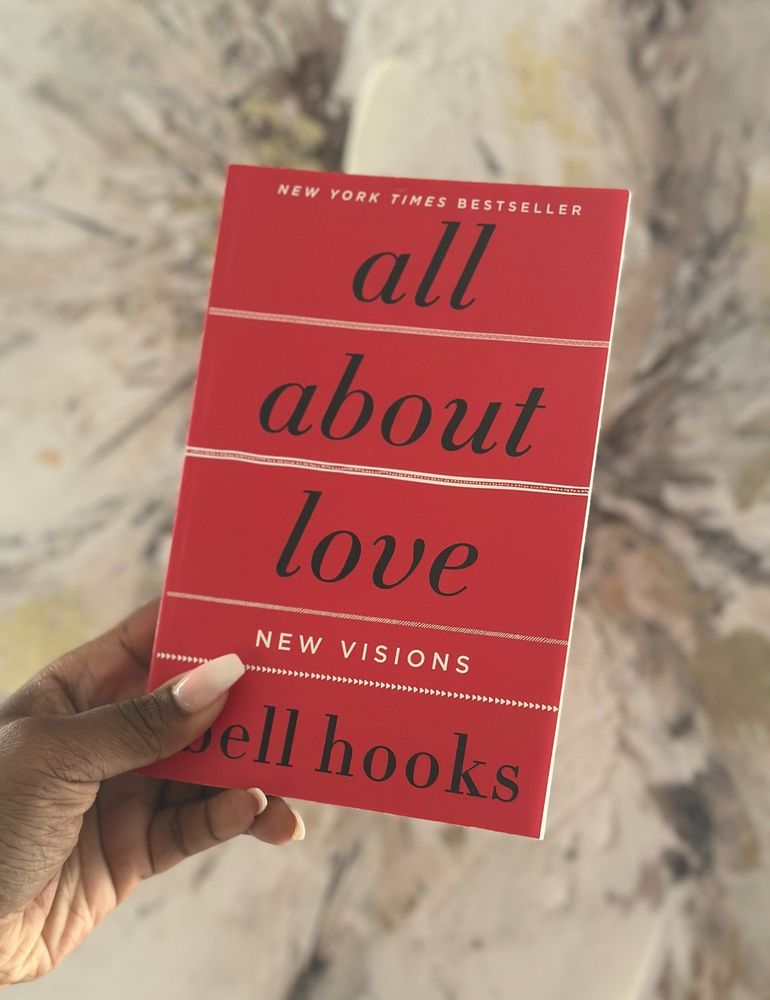 The image shows a hand holding a copy of the book All About Love: New Visions by bell hooks. The cover is a bold red with black, lowercase text for the title and author’s name. The title is presented in a simple, elegant font, and the book is noted as a New York Times Bestseller. The background is blurred, featuring a soft, marbled pattern in neutral tones. The hand has light-colored nails and is holding the book from the lower-left corner.
