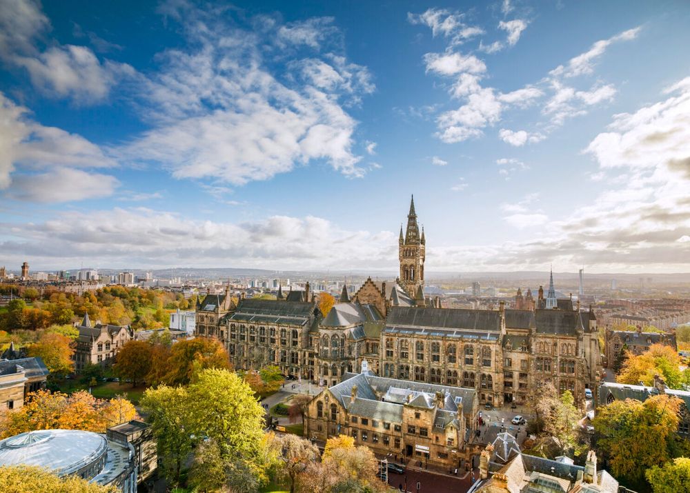 University of Glasgow main building with blue skies and fluffy white clouds in the backdrop. The trees have green and orange leaves and the city is in the background. 