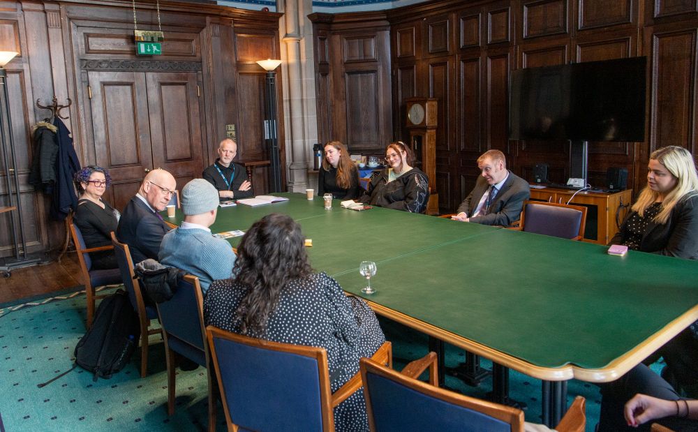 The group of UofG students and colleagues sitting round a table talking with John Swinney.