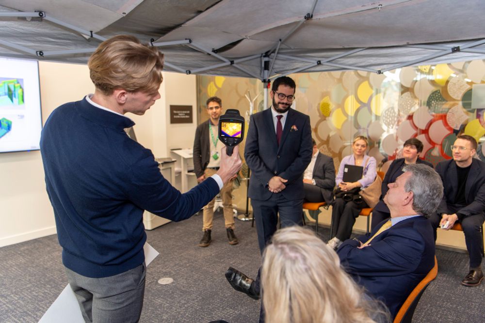 A man demonstrates a thermal imaging device to a group of people under a canopy indoors.