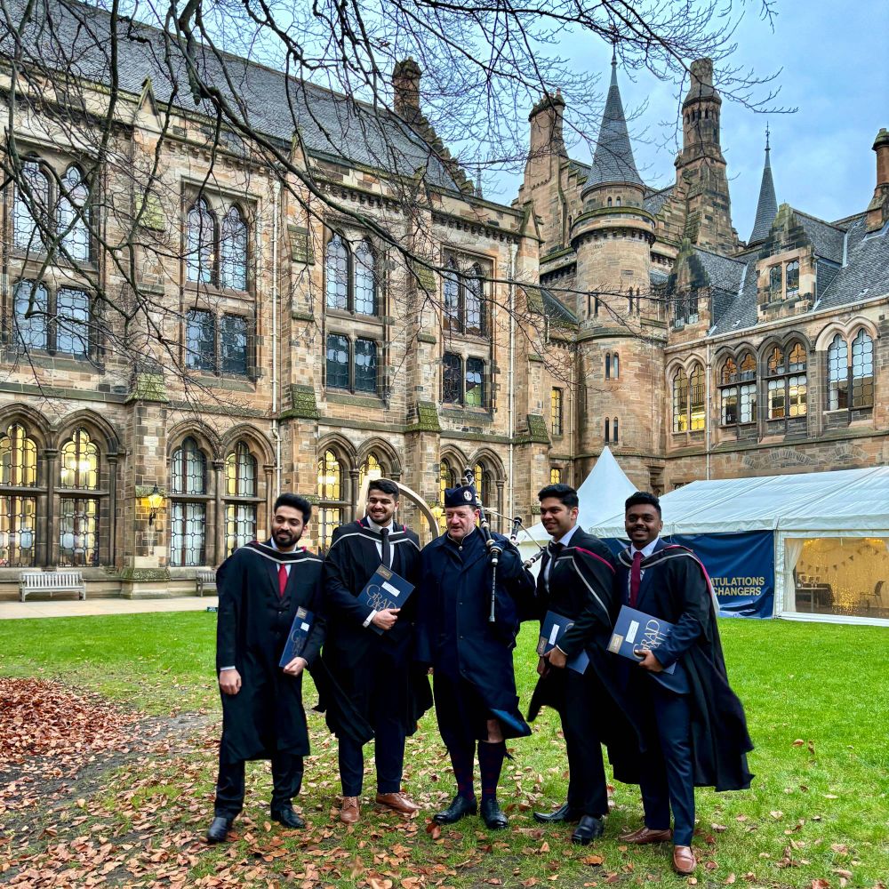  A group of graduates in academic gowns posing outdoors on a lawn in front of the University of Glasgow's iconic stone building. A bagpiper in traditional Scottish attire stands with them, holding a set of bagpipes.