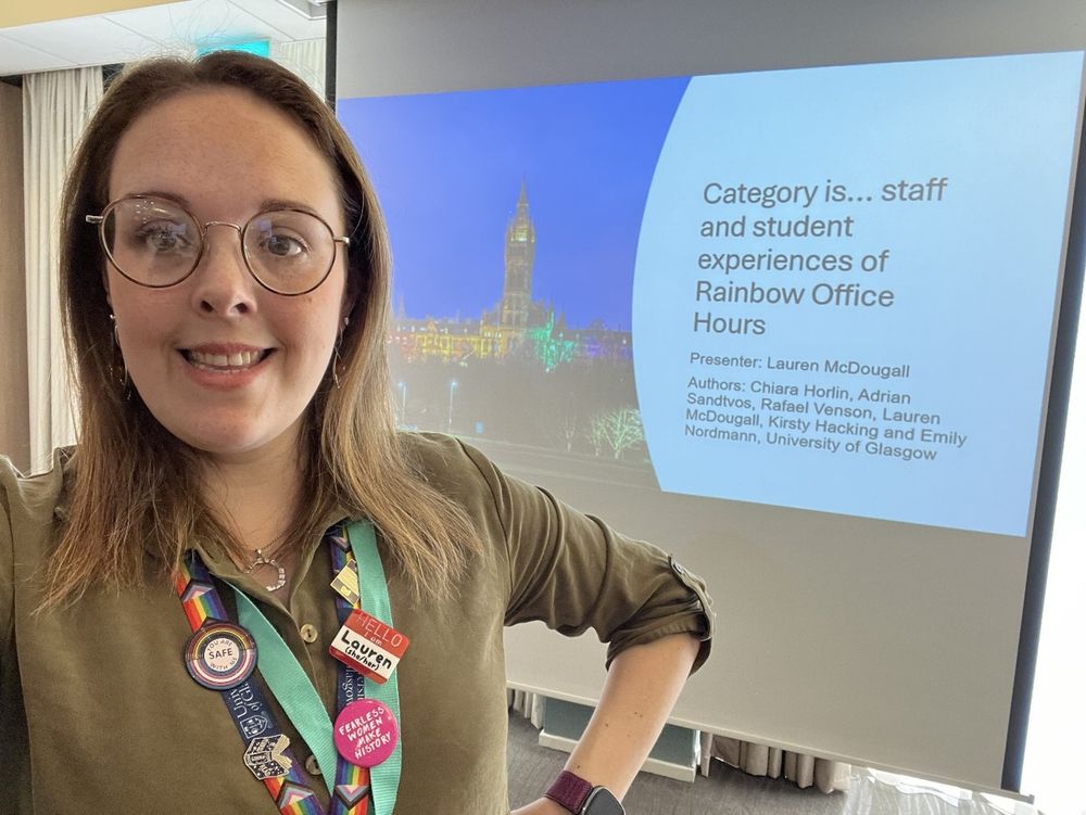 Lauren stands in front of a presentation screen, it's titled: Category is... staff and student experiences of rainbow office hours. 
