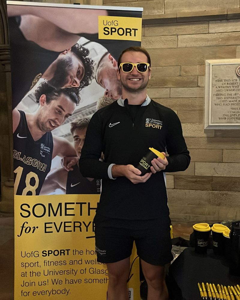 A photo of a member of staff from UofG Sport holding a water bottle, smiling, next to a pop up banner