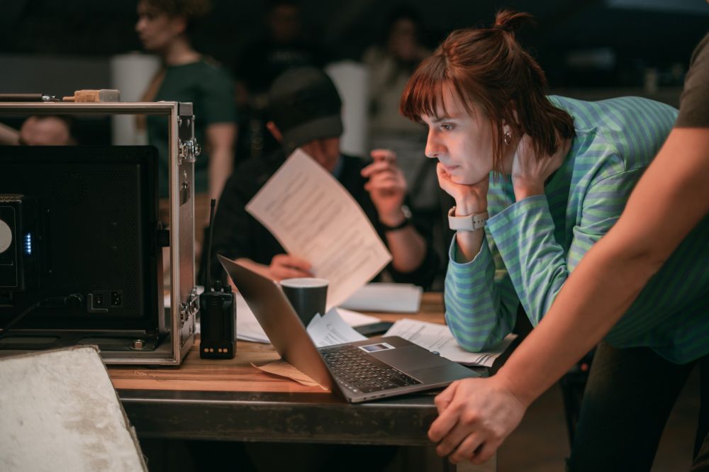 A woman stands over a laptop with a group of people in the background around her on set. 