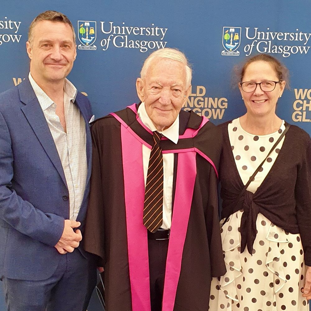 Dr Michael Simpson wearing graduation robes and standing with his family in front of a banner which reads 'World Changing Glasgow'. 