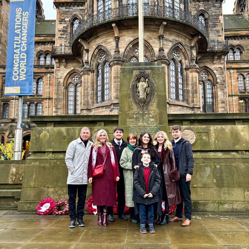 A family group gathered in front of a monument at the University of Glasgow. The backdrop  features stone architecture with a banner reading "Congratulations World Changers."