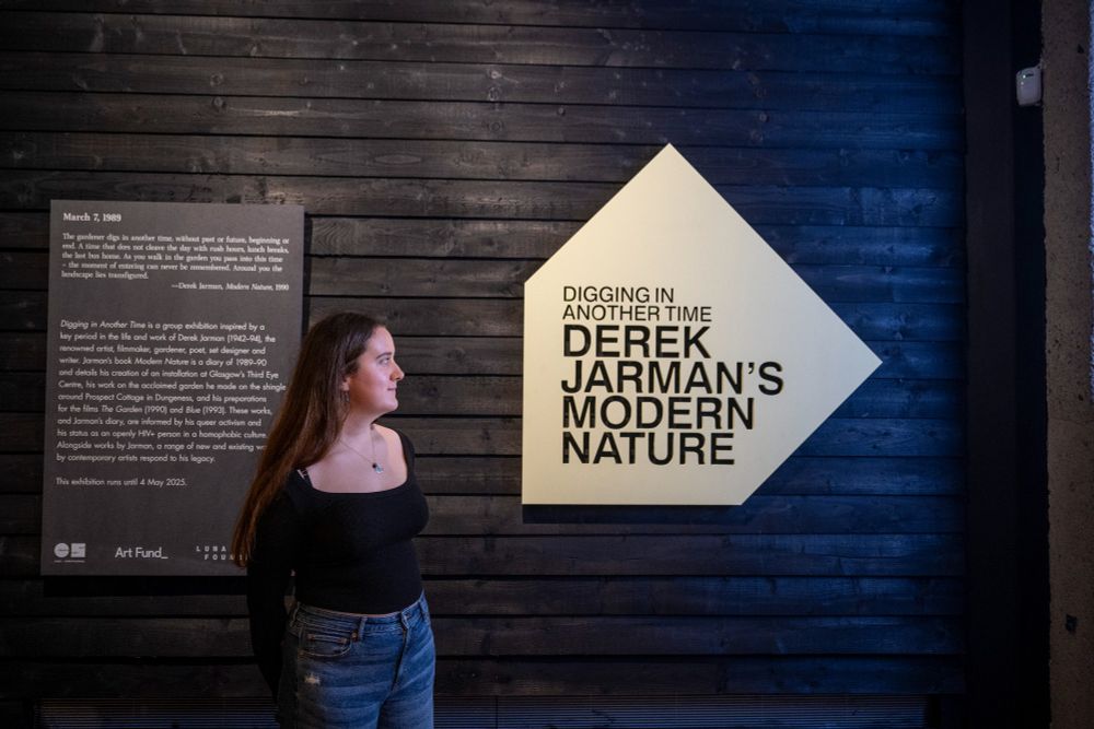 A woman stands in front of a dark wooden wall with a large yellow sign reading "DIGGING IN ANOTHER TIME: DEREK JARMAN'S MODERN NATURE." A plaque to the left provides historical context on Derek Jarman’s work and activism.