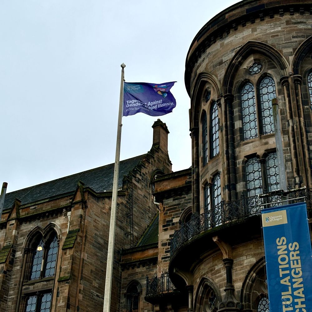 A purple flag with the words Together Against Gender-Based Violence written on it, is blowing in the wind. It is flying outside the gothic main building at the University of Glasgow.