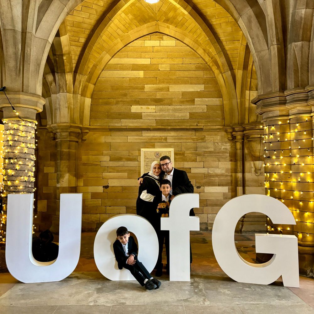 A family of four poses under an archway in the University of Glasgow cloisters with large white letters spelling "UofG." The graduate is in academic attire, and one of the children is sitting in the 'O' of 'UofG'.
