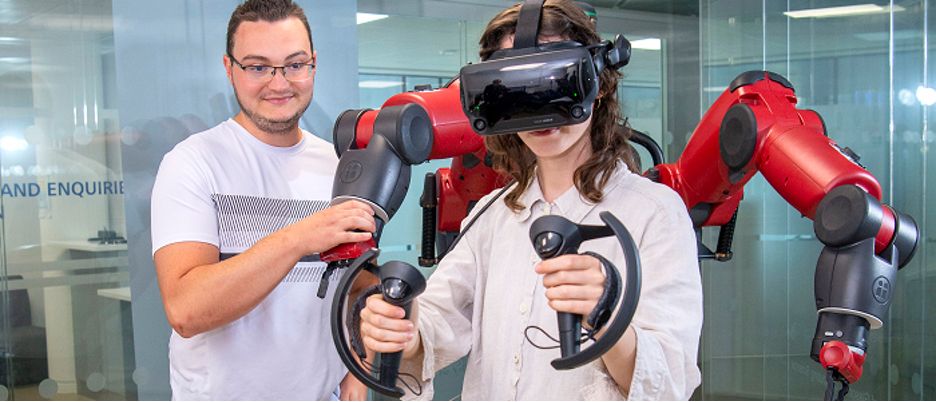 A woman wearing a virtual reality headset and holding VR controllers interacts with a red robotic system. A man beside her observes and adjusts the robot’s arm. They are in a modern lab or office environment with glass walls.