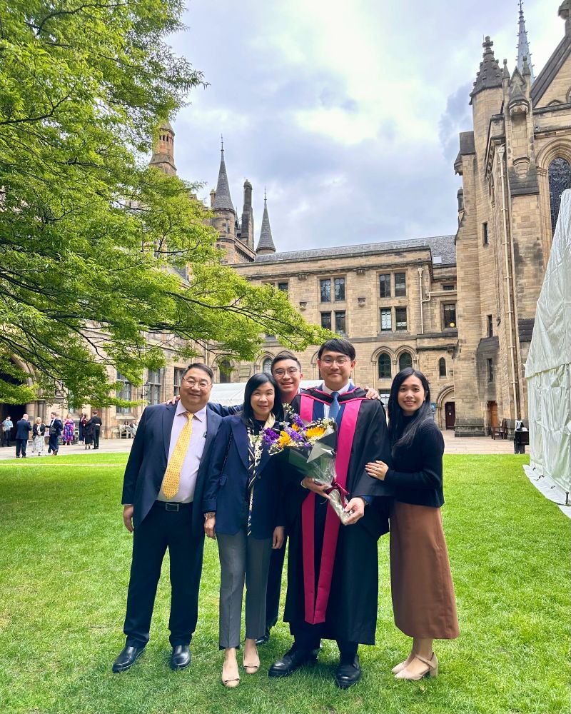 A graduate wearing robes and holding flowers standing the in the quad with his family.