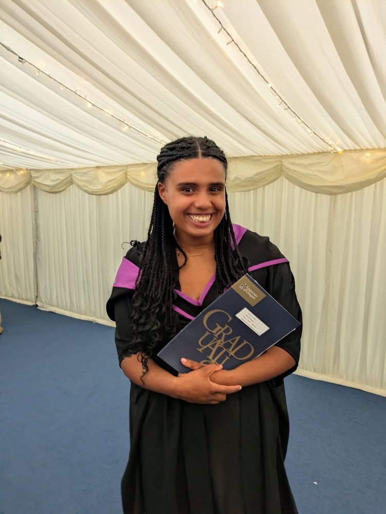 A graduate in a tent holding a blue folder with the word 'Graduation' written in gold.