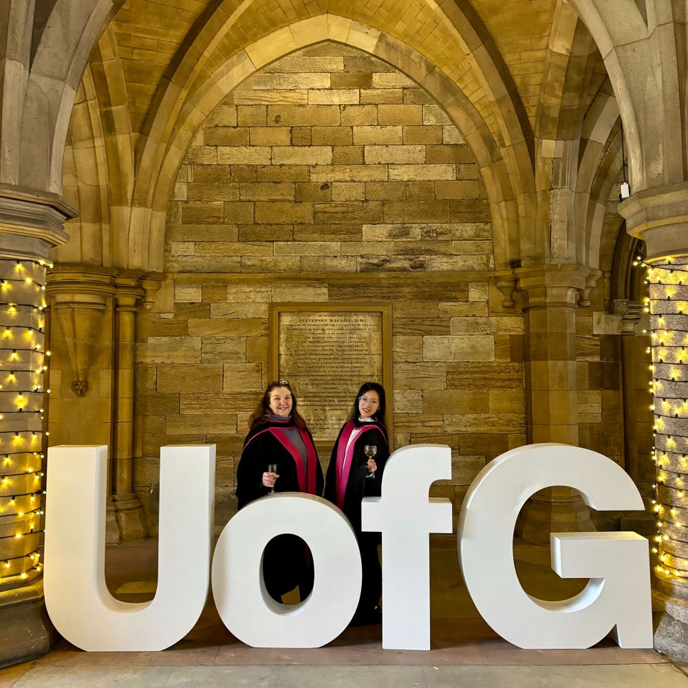 A graduates and a academic colleague wearing academic gowns pose with champagne flutes next to large white letters spelling "UofG," set in an arched stone hallway with soft decorative lighting.