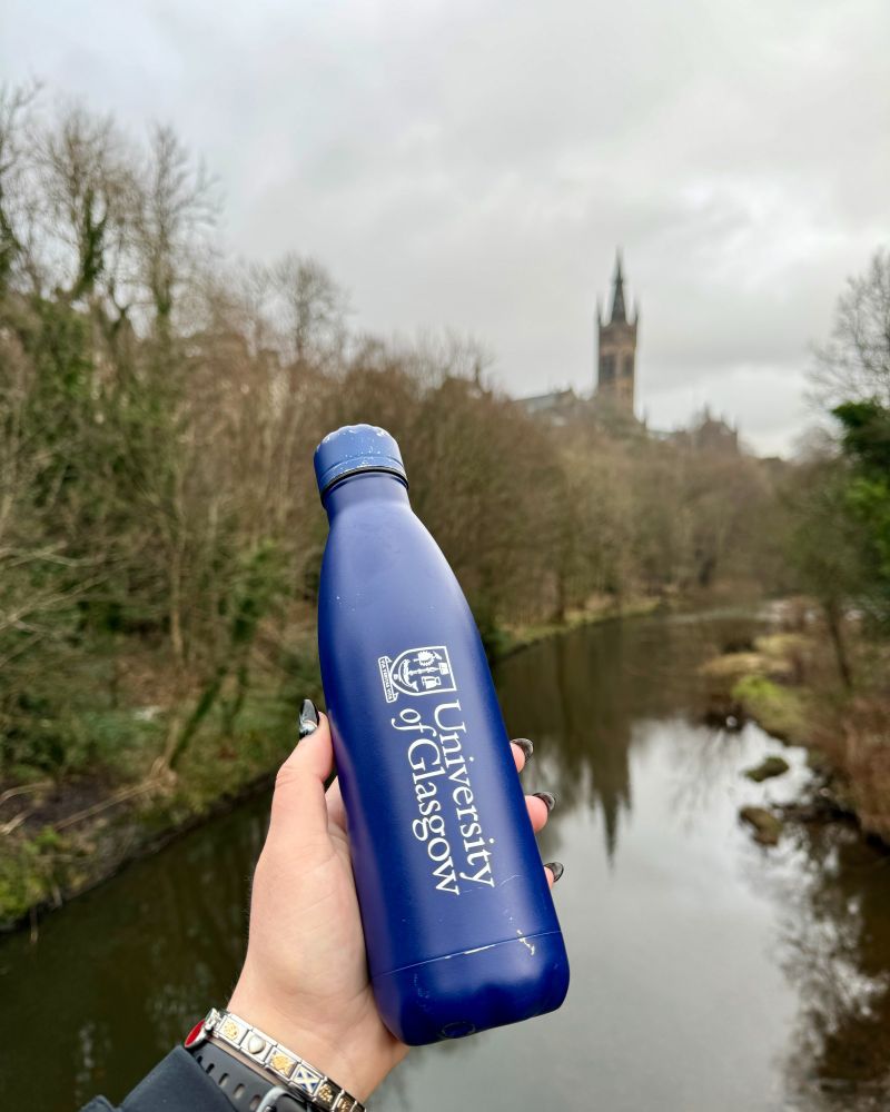 A woman holding a reusable blue hot drink bottle in their hand with the University of Glasgow white logo on it. The River Kelvin, Kelvingrove Park and the University of Glasgow are in the background, with the Tower of the main building reflecting in the river.