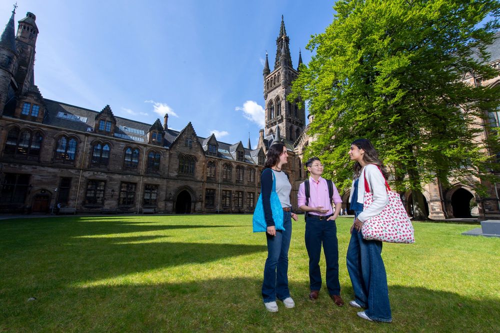 Three student standing in the university quads on a sunny day, behind them in the tower and Gilbert Scott building. 