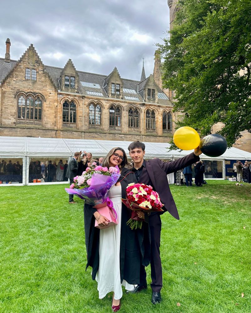 A graduate holding two bunches of flowers standing with another person holding gold and black balloons. 