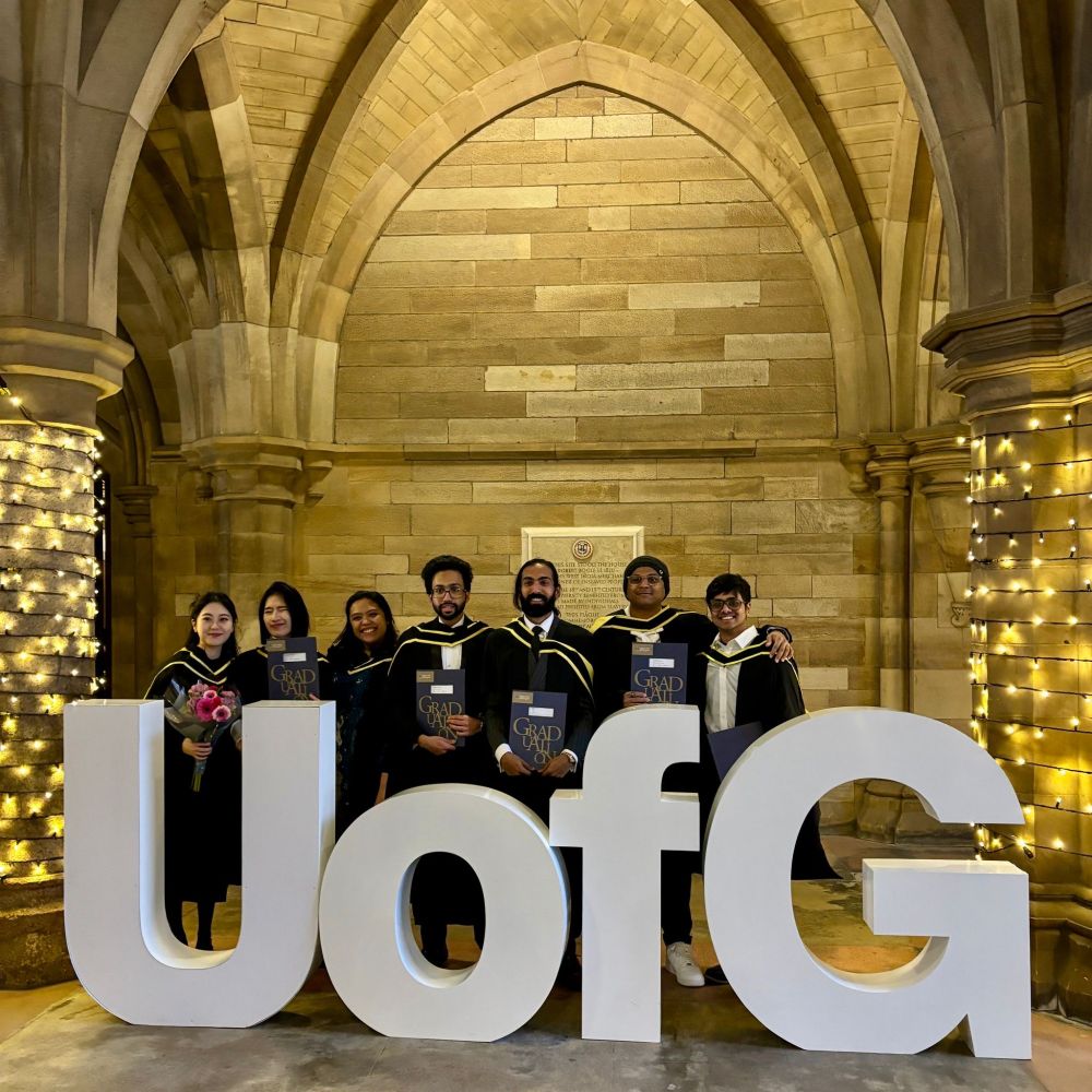 A group of graduates in academic gowns and hoods pose together under an stone archway in the cloisters with fairy lights wrapped around the columns. They are holding their diplomas and standing behind large white "UofG" letters.