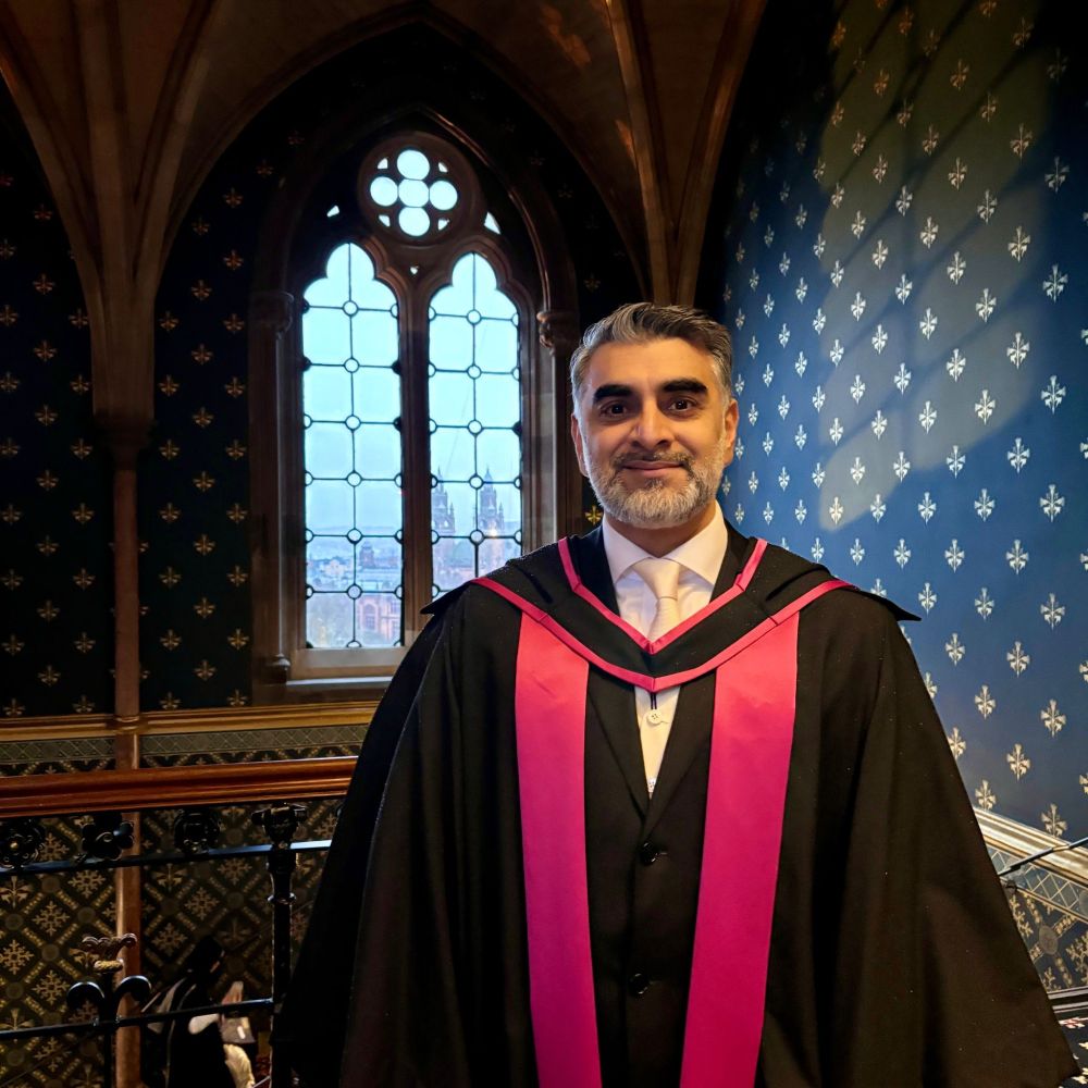 A graduate in a black academic gown with a pink hood stands inside the historic Gilbert Scott building. There is patterned wallpaper and a large Gothic-style arched window in the background. 