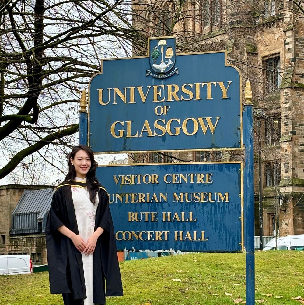 A smiling graduate in traditional academic gown and hood stands in front of the blue and gold University of Glasgow sign. The background includes trees and the historic campus main building.