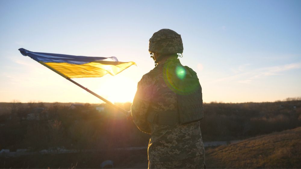 A soldier in camouflage military uniform and helmet stands on a hill at sunset, holding a waving Ukrainian flag. 