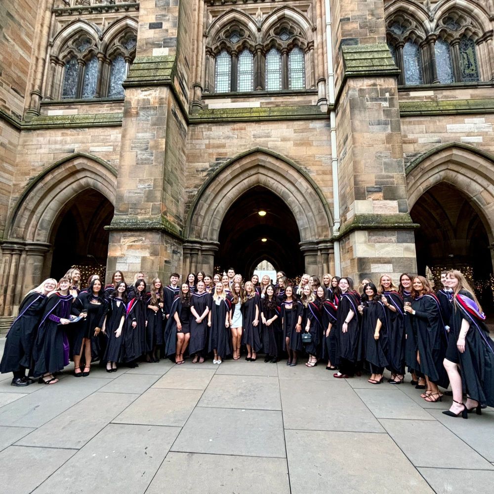 A big group of graduates in academic gowns standing together outside the university cloisters which feature tall arches and intricate stonework. They are smiling and posing for a group photo on a paved courtyard.