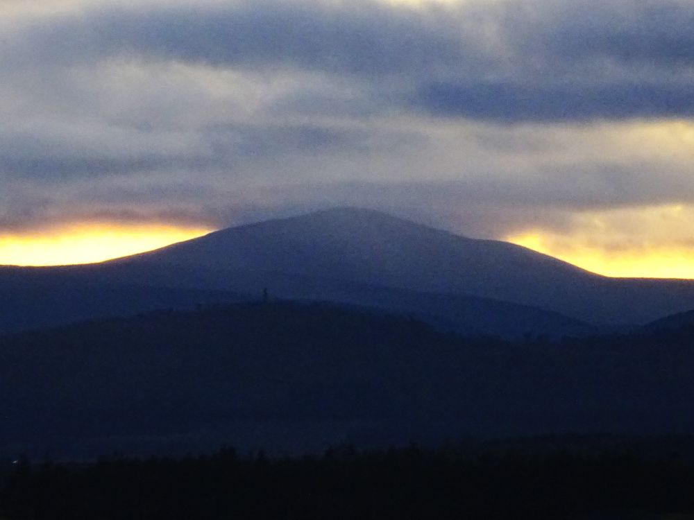 A landscape photo after sunset with just a band of gold behind the hills. Closest to the viewer the land is black, it lightens with each layer of distance. A hill in the mid distance has a tower on it, a tiny dark line in the photo. Grey blue cloud is pressing down on the top of the furthest hill.