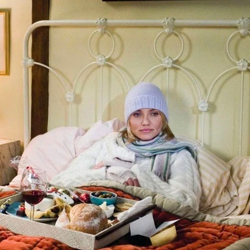 A photo from the movie The Holiday where Cameron Diaz’s character is sitting in bed, wrapped in a hat and scarf, with a tray of wine, cheese, bread, and other snacks on her lap.