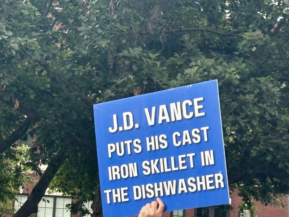 A blue sign that says “JD Vance puts his cast iron skillet in the dishwasher” with trees in the background.