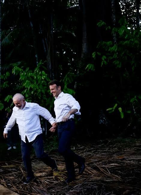 Lula e Macron de mãos dadas correndo próximo a algumas árvores. Ambos usam camisas brancas. Eles riem juntos