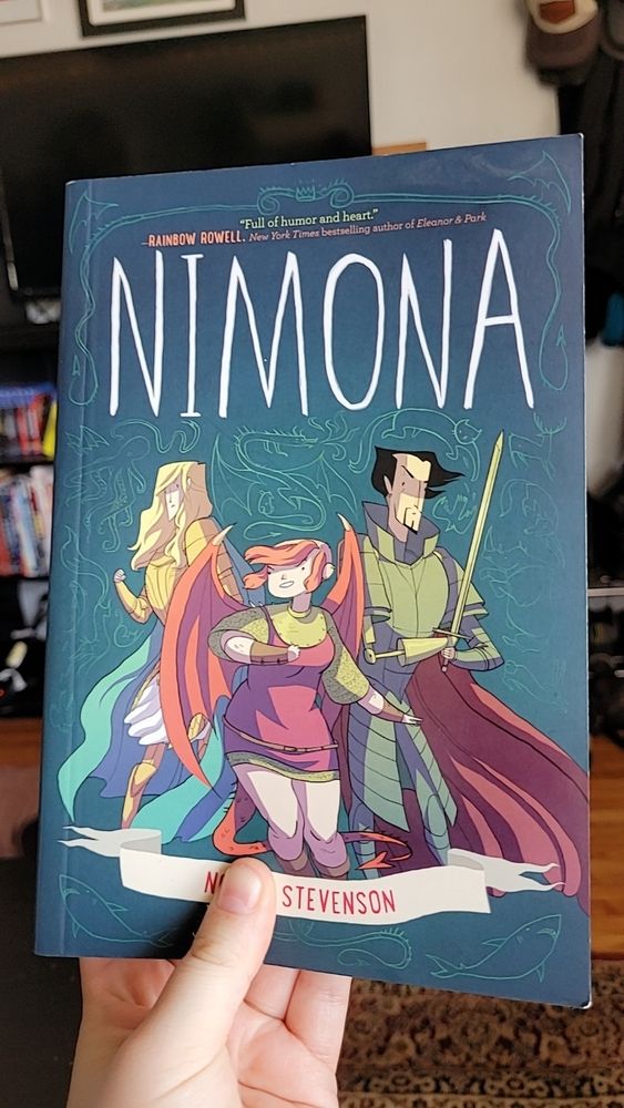 A comic book held up by hand, showing the cover of 'NIMONA', three fantasy characters on a green background. On the left is a blond man in golden armor, on the left is a dark haired man with sharp facial hair in armor and a red cloak. In the center is the title character, a red haired punk style girl in chain mail with red dragon wings. The author is N. Stevenson. 