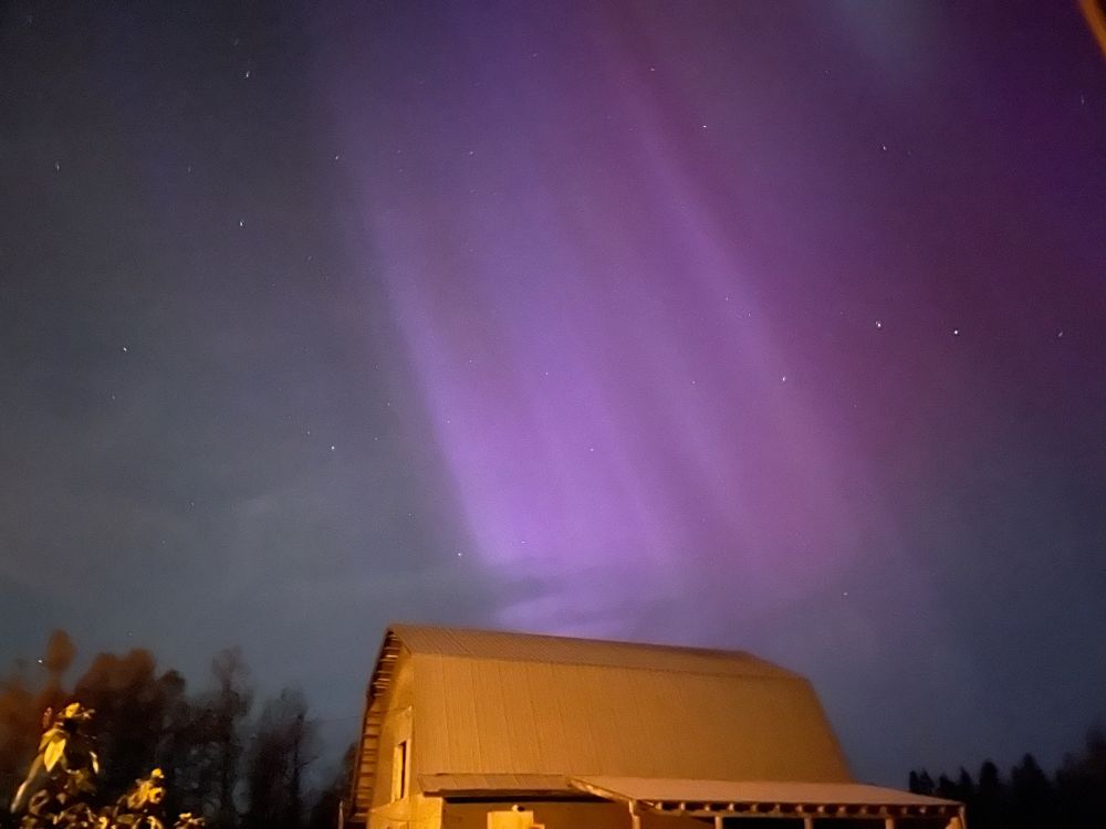 The northern lights are captured in a purple hue against the night sky. In the foreground, a barn is illuminated by floodlights.