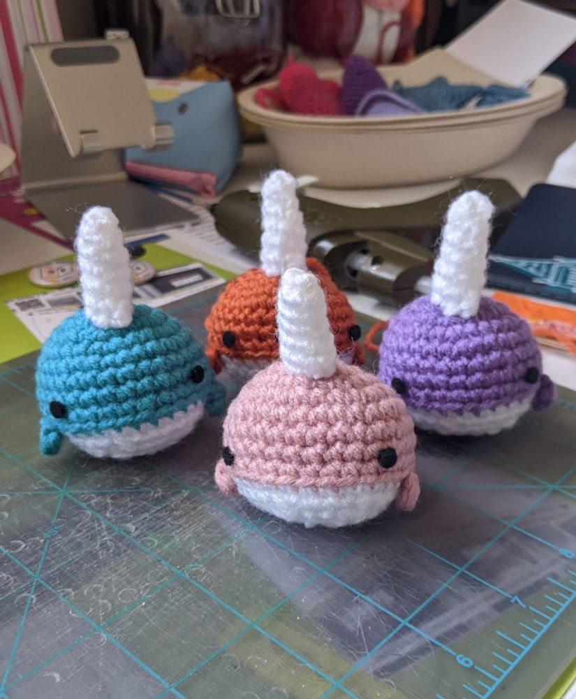Four crocheted narwhal stuffies in colors of pink, lavender, red orange, and aqua. 