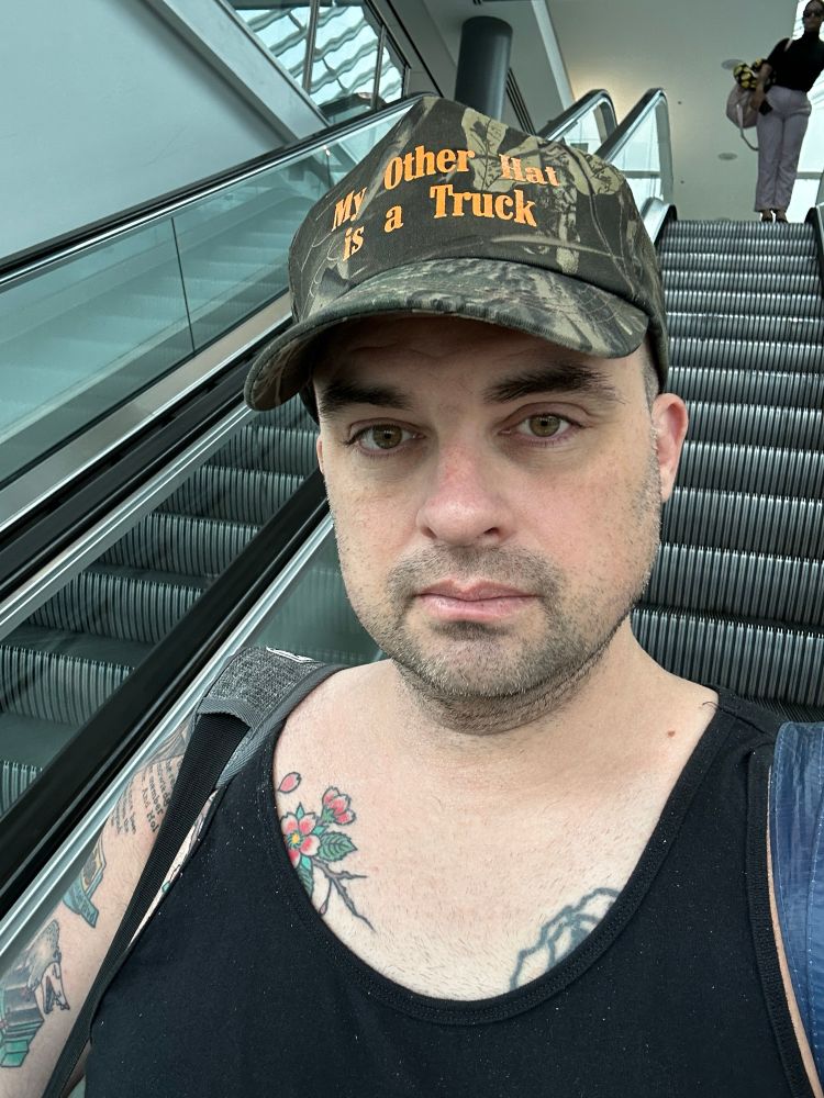 A gay man wearing a camo hat with text that says “my other hat is a truck” 