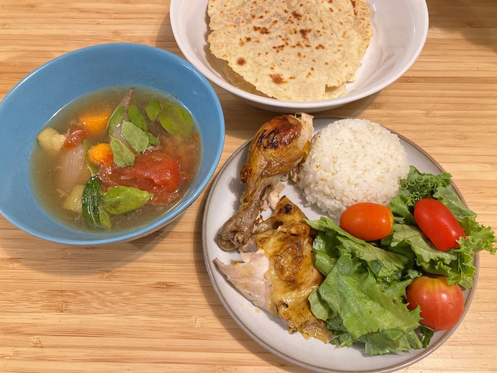 A bowl of soup with vegetables, next to a plate of chicken, rice, salad, and another plate of flat bread. 