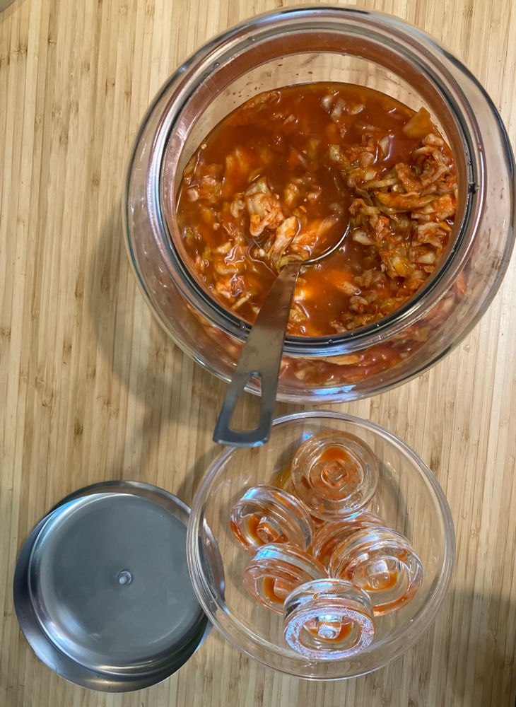 A 7.57L (2 gallon) jar half filled with fresh kimchi, a bowl of fermentation weights to one side.
