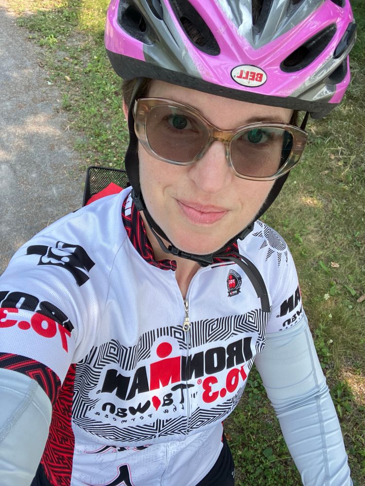 A selfie of a middle aged white woman mid-ride on a gravel trail. She has short hair, glasses, and is wearing a helmet. 