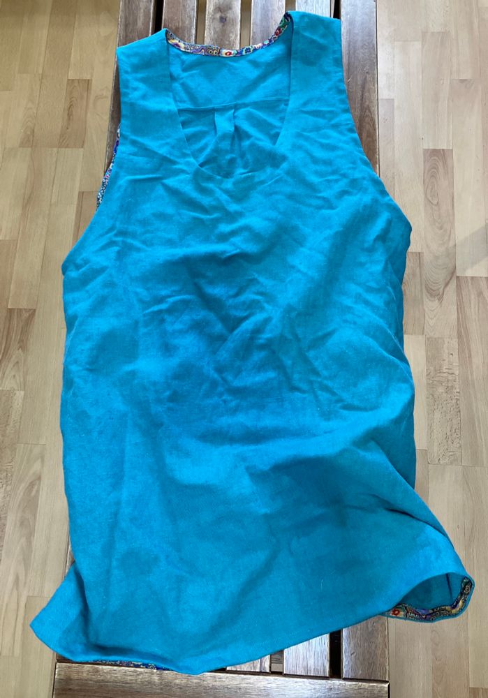 A teal linen tunic laid flat on a bench with colourful print bias binding on the neck and armholes and the hem.
