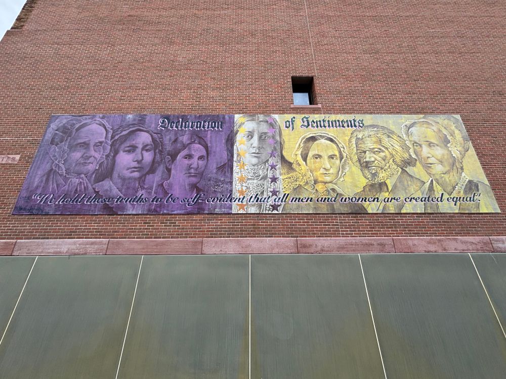 Mural at Women’s Rights National Park. Half is purple, half is yellow/good. Images of women and Frederick Douglas from the shoulders up are in the background. The top reads “Declaration of Sentiments.” The bottom reads “We hold these truths to be self-evident that all men and women are created equal.”