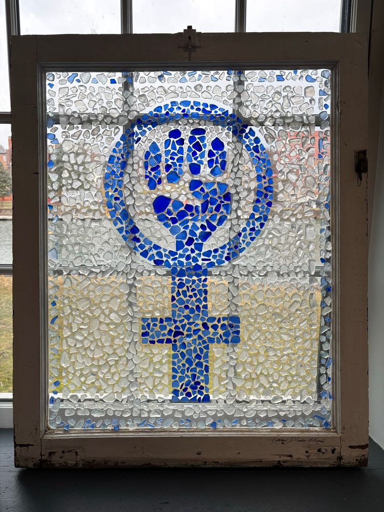 Stained glass image of the female symbol with a fist, or the feminist symbol. The symbol is blue and the surrounding area in the frame is clear. The glass is made out of pebbles rather than shards. Found at the National Women’s Hall of Fame. 