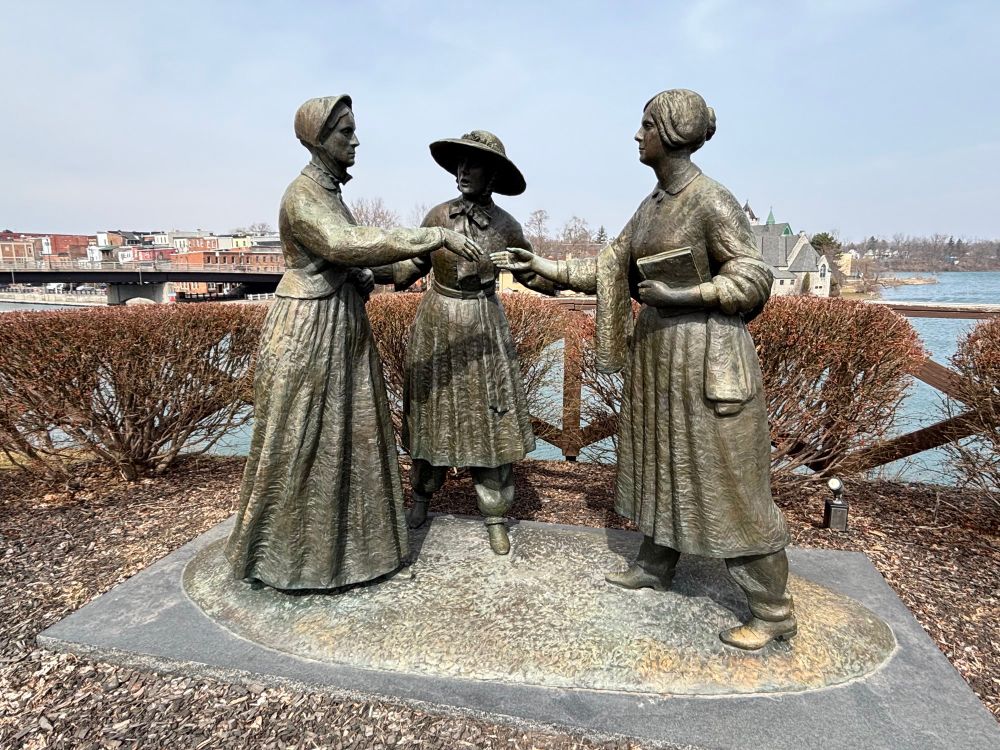 The statue “When Stanton Met Anthony” found in Seneca Falls, NY. Three female figures are standing in 19th century clothing. They are life-sized bronzed sculptures. Susan B Anthony is on the left, Amelia Bloomer is in the center, Elizabeth Cady Stanton is on the right. Anthony’s hand is outreached to grasp Stanton’s hand. Bloomer’s arms are outreached to both. 