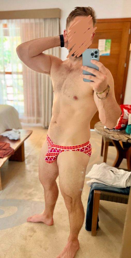 Standing with one hand behind my head in a brightly patterned bikini brief swimsuit with a prominent bulge in a Mexican hotel room. The geometric design on the bikini accentuates my package. My body is lean and muscular with trimmed body hair. 