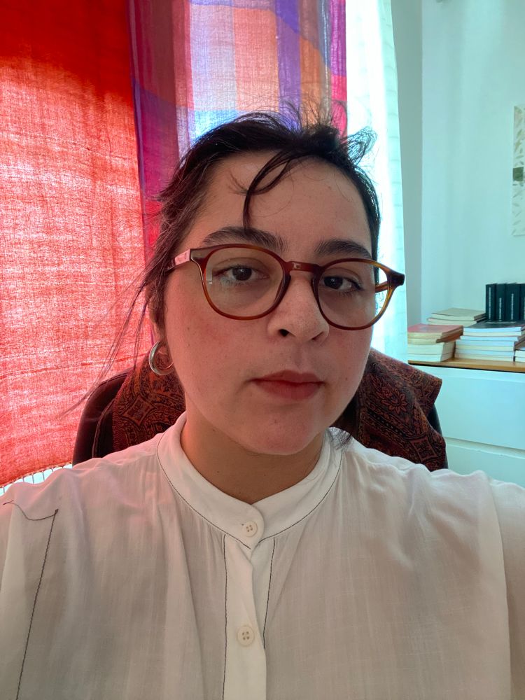 selfie de uma mulher branca de cabelos pretos sentada em uma poltrona. ao fundo lenços alaranjados e uma cômoda branca com livros em cima 