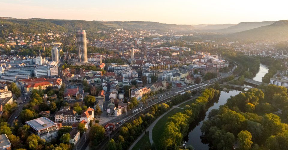 A medium sized German city. On the right is a picteruesque river in the glowing daybreak. Around the city are gently rolling, green hills.