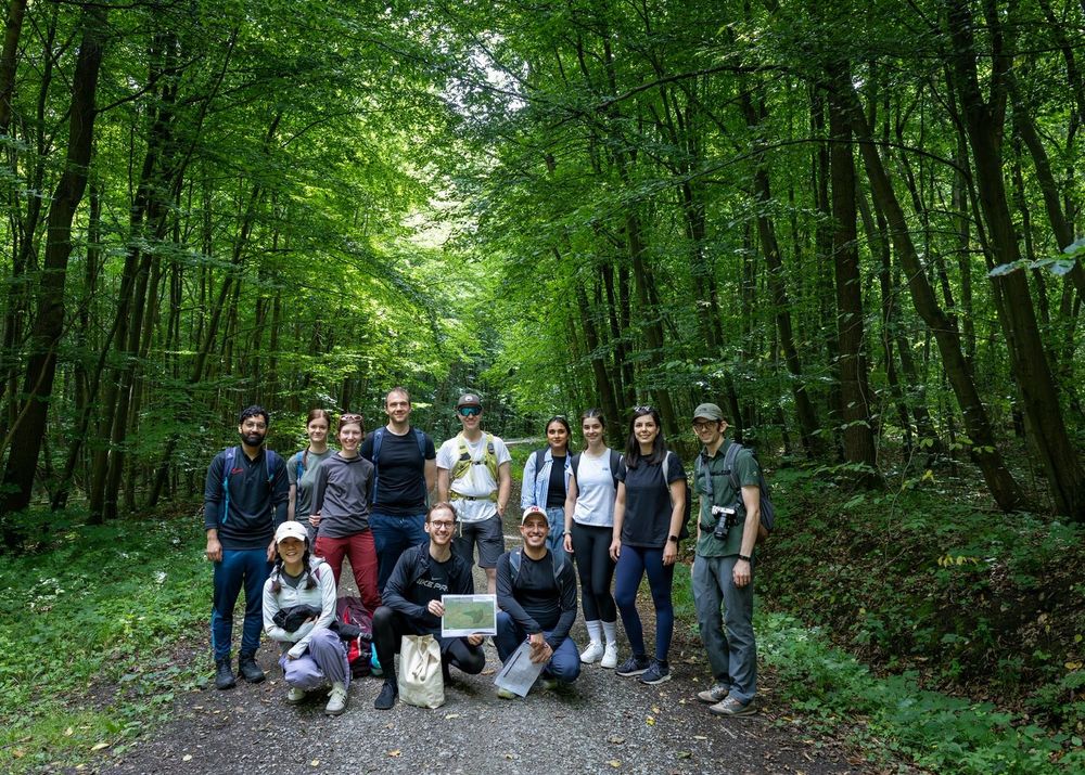 Two rows of people wearing active or outdoors clothing stand in a lush green forest. One person in the front row holds a map of the area.