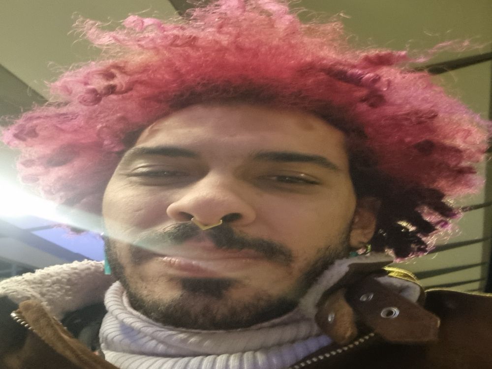 A selfie of Edwin, a slender, brown-skinned person with a red and pink-ish afro and a black beard gazing with a tired but satisfied expression into the camera. The light beam of a lamp is shining brightly into the lens from the side. Behind them the roof of one of the tracks of the Basel station can be seen. 