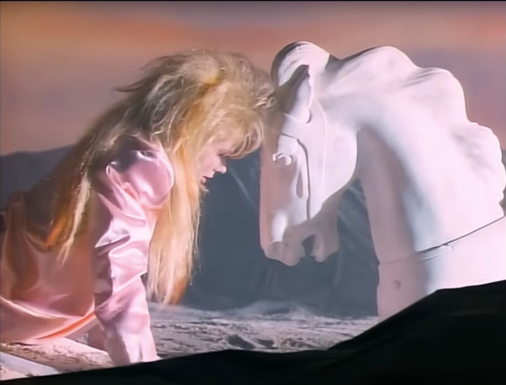 Nancy Wilson in the Heart video for These Dreams, in a pink jacket with rocker hair, forehead (hair) touching the forehead of a white knight chesspiece in the desert, pastel sky beyond
